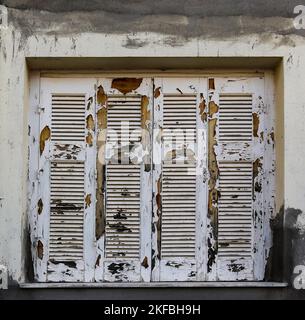 Extremely chipped and weather-worn white paint on closed grungy wooden shutters in stucco building with grey paint smeared abound Stock Photo