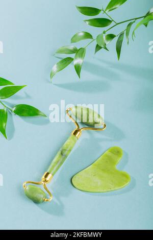 Green jade face roller and gua sha scraper with leaves on light blue background Stock Photo