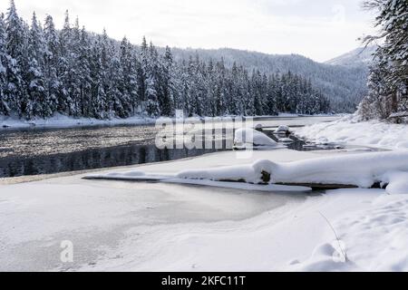 Snow-Covered Trees and Rocks Along Partially Frozen Lake in Winter Stock Photo