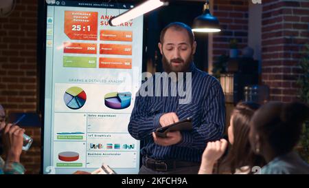 Company analyst explaining statistics diagrams, giving presentation in business meeting, holding digital tablet. Employee showing analytics research charts on screen at night time. Handheld shot. Stock Photo