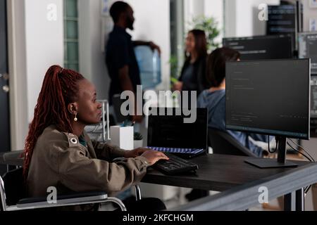 Programmer in wheelchiar typing html code on computer, working with cloud computing terminal window. Disabled software developer writing artificial intelligence algorithm in it company office. Stock Photo