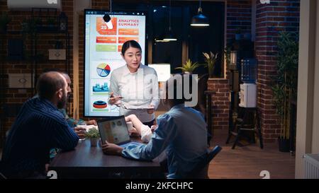 Asian company analyst explaining team revenue statistics in business meeting, financial report presentation, employee holding clipboard. Diverse coworkers discussing analytics data, talking Stock Photo