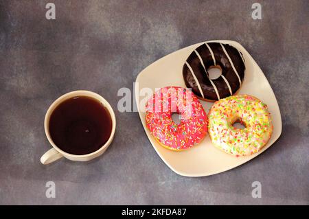 A plate with three donuts in multi-colored glaze and a cup of black coffee on a gray abstract background. Top view, flat lay. Stock Photo