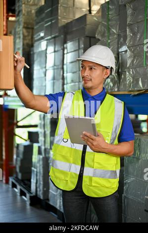 Portrait, Handsome smart millennial Asian male warehouse supervisor worker in his uniform working in warehouse, using tablet, checking inventory. Carg Stock Photo