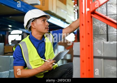 Concentrated and professional Asian male warehouse worker or foreman in white hardhat and uniform, checking an inventory, working in warehouse. Stock Photo