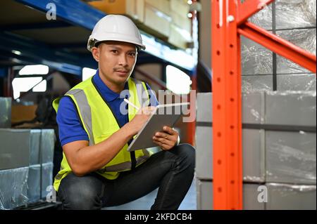 Handsome and professional Asian male warehouse worker or foreman in white hardhat and uniform, checking an inventory list on tablet, working in wareho Stock Photo