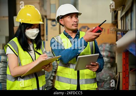 Professional adult Asian male warehouse supervisor training a new female worker, checking their inventory in the warehouse or storehouse. Stock Photo