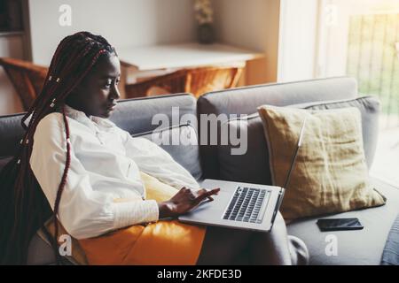 View of a thoughtful young black woman entrepreneur in a white shirt sitting on the sofa of her domestic living room and using a netbook; black female Stock Photo