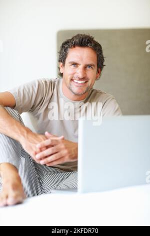 Happy man using laptop in bed. Portrait of handsome man sitting with laptop on bed. Stock Photo