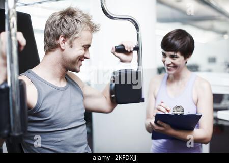 How am I doing, coach. a handsome young man using an exercise machine while his personal trainer looks on. Stock Photo