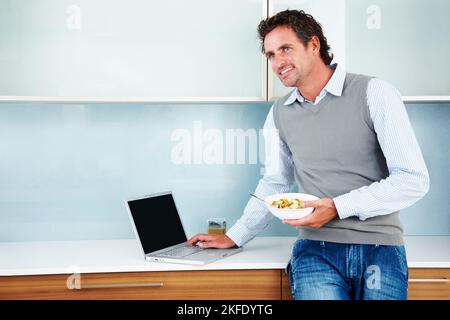 Confident mature man using laptop in kitchen. Confident mature man using laptop while holding fruit salad in the kitchen. Stock Photo