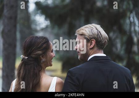 Rear view portrait of adorable just married couple. Turkish middle-aged long-haired woman and her new husband - Scandinavian blond bearded man - standing arm to arm and looking at each other. Blurred forest trees in the background. High quality photo Stock Photo