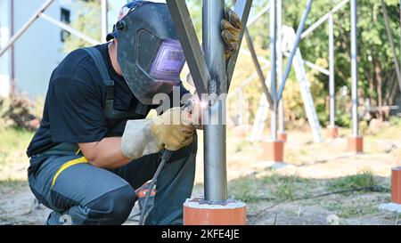 Welder welding metal beam with welding machine for installing solar panels. Concept of renewable and ecological energy. Idea of environment safe. Man in protective welding helmet wearing workwear Stock Photo
