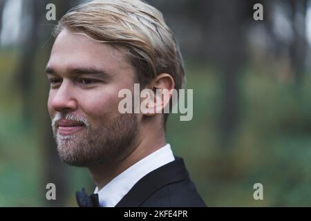 Wedding memories. Closeup portrait of handsome elegant Scandinavian blond bearded groom in black suit with butterfly tie looking far ahead. Blurred background. Forest photoshoot. High quality photo Stock Photo