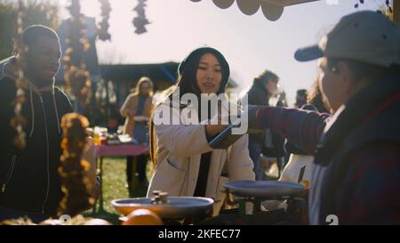 Senior farmer gives paper bag with goods to African American man. Asian woman contactless pays for purchases using smartphone. Shopping at local farmers market on weekend. Vegetarian and organic food. Stock Photo