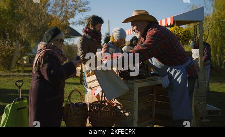 Senior farmer selling fruits and vegetables at the stall. Elderly woman puts products in paper eco bag. Local farmers market or autumn fair outdoors. Agriculture. Point of sale system. Stock Photo