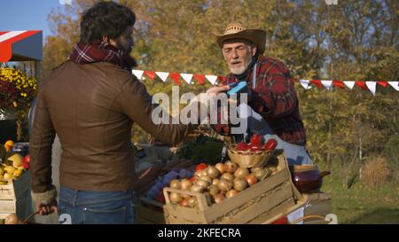 Adult man contactless pays for purchases to farmer using smartphone. Weekend shopping at local farmers market outdoors. Autumn fair. Vegetarian and organic food. Agriculture. Point of sale system. Stock Photo