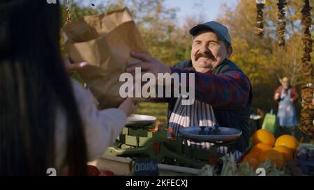 Senior farmer seller weighs bananas on scale, puts them in paper eco bag and gives to woman. Outdoors shopping at local farmers market on weekend. Vegetarian and organic food. Agriculture. Stock Photo