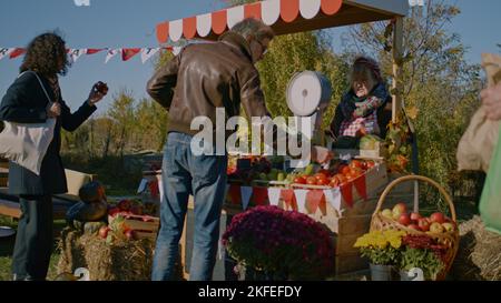 Mature man in sunglasses choose fresh vegetables or fruits and weight them. Holder of sales point offers own products. Shopping at local farmers market on weekend. Vegetarian and organic food. Stock Photo