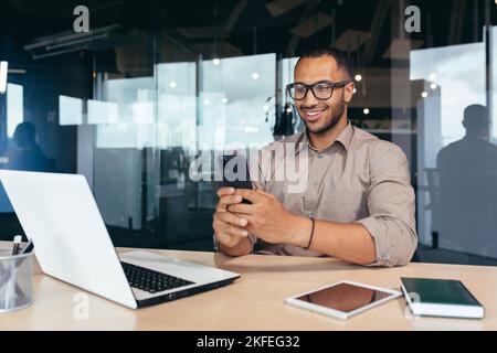 Happy and smiling businessman rejoicing at successful work results and achievement, man at work using phone reading online good news sitting at table inside office, using laptop at work. Stock Photo
