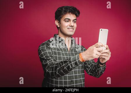 A smiling young Indian man in a checkered shirt takes a selfie on a smartphone against a red background. On a video call with a girlfriend or fiancee. Stock Photo
