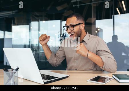 Successful programmer in office with laptop celebrating successful completion of software project, startuper in overcoat holding hands up gesture of triumph and achievement. Stock Photo