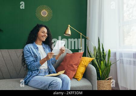 Happy young red-haired Hispanic woman received a letter. Sitting at home on the sofa. Holding a letter with an envelope, reading, smiling. Stock Photo