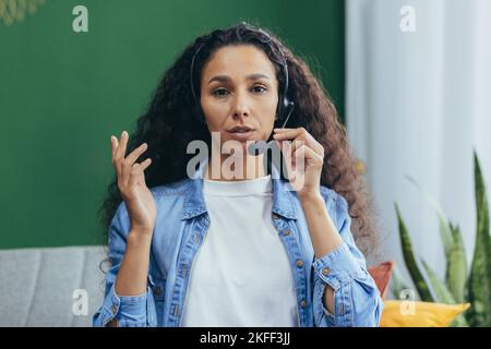 Close-up photo. Young beautiful Latin American woman talking on video call online into the camera wearing headphones with a microphone. Stock Photo