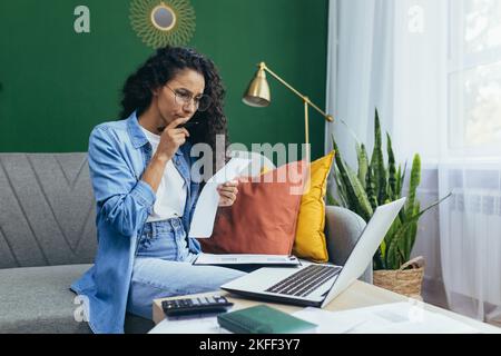 Concentrated and serious young beautiful Hispanic businesswoman, freelancer working at home on laptop, holding documents in hands, sitting on sofa. Stock Photo