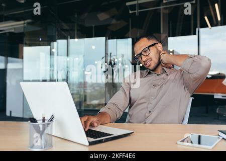 Office worker has severe neck pain, businessman in glasses working inside office using laptop at work, african american executive overtired. Stock Photo