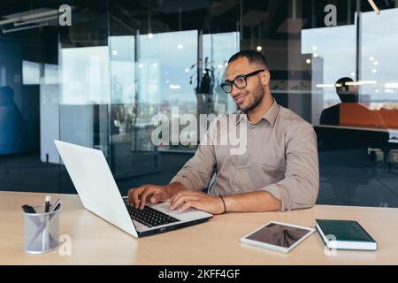 African american programmer at work with laptop inside office, man in glasses and casual writing code for programs. Stock Photo
