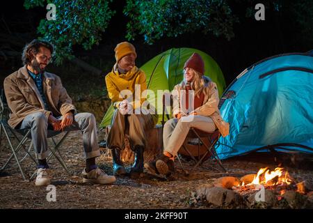Three happy friends enjoying camping with tent chatting by a fire having fun together in nature Stock Photo