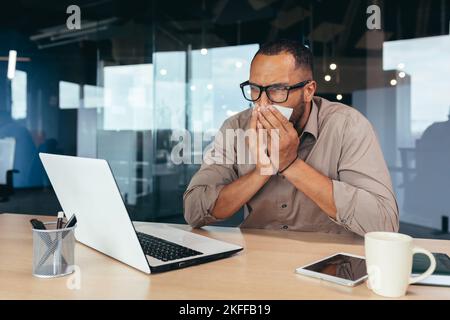 Man sneezes and has runny nose in office at work african american businessman sick working inside office at desk using laptop. Stock Photo