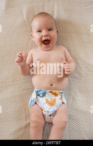 Happy baby girl lying on her back on a bed. She is wearing a modern, reusable cloth nappy with a pretty print. Stock Photo