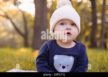 baby in white knitted hat sits on grass in Park against background of autumn trees Stock Photo