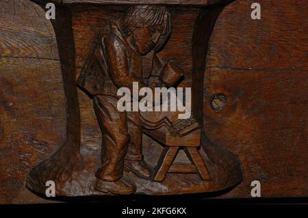 Dutch sculptor Joop van Huisstede (1922-2000) immortalised his son Koos in this sculpted misericord he carved in 1974 for the Oude Kerk (Old Church) in Ouderkerksplein, Amsterdam, Netherlands.  The artwork, which joined late-15th century carvings beneath the choir stall seats, shows the young man working on one of the partitions between the stalls. Stock Photo