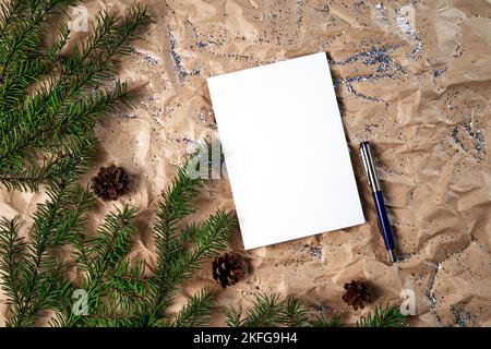 Empty white notebook and pen on crumpled kraft paper Christmas background with fir branches, mock up. Happy New Year and Merry Christmas. New Year's goals or promises with colorful decorations on the background of crumpled kraft paper with Christmas tree branches. High angle. Stock Photo