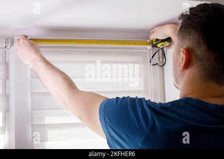 A handyman with a tape measure installs white roller blinds indoors. Shutters day and night. Stock Photo