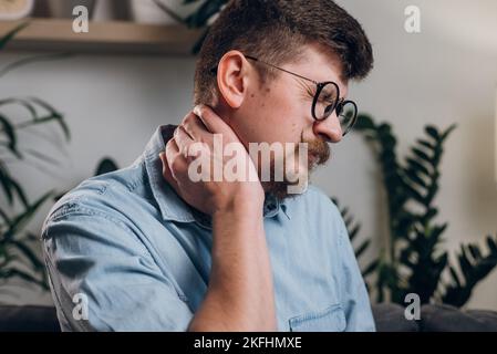 Young bearded man in eyeglasses studying until night at computer with neck pain. Close up of tired worker kneading neck, muscle recovery and massage. Stock Photo
