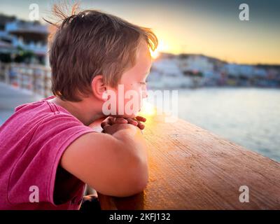 Side view of thoughtful cute boy looking at sea while resting head on wooden railing against sky during sunset Stock Photo