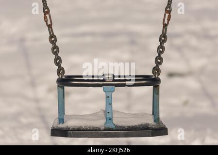 Old blue child's swing with snow in the background. Cold winter day in the play ground. Rusting chain links. Stock Photo