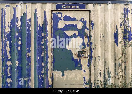 Rustic metal unit with the word canteen on a sign above the door. Blue paint peeling off to reveal white and green rusty paint. weeds growing at the s Stock Photo