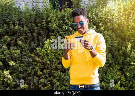 A smiling young African man wearing a yellow sweatshirt and sunglasses holds his cell phone in one hand and a payment card in the other.