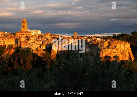 View across olive-branches to medieval town Pitigliano glowing in evening light with cathedral Santi Pietro e Paolo and tower in the centre, Maremma, Stock Photo