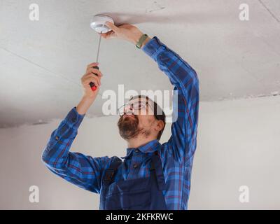 young electrician man in blue work suit installing smoke detector on the ceiling Stock Photo