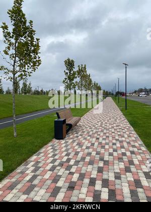 walking path in the park is paved with multi-colored paving stones. Bench and trash can next to the sidewalk. Bicycle path in the background. Selectiv Stock Photo