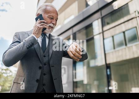 Senior businessman checking time on wristwatch. Male professional wearing suit is talking on smart phone. He is standing against corporate building of Stock Photo