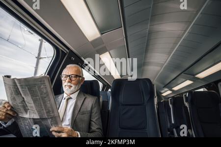 Senior entrepreneur reading newspaper while sitting by the window. Male professional is traveling in modern passenger train. He is wearing suit while Stock Photo