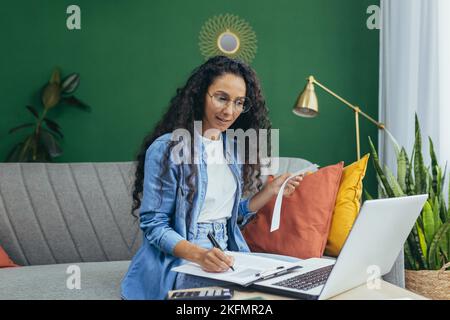 Young beautiful hispanic woman student studying online at home from laptop. Sits on the sofa, writes, listens to a lecture, passes an exam. Stock Photo