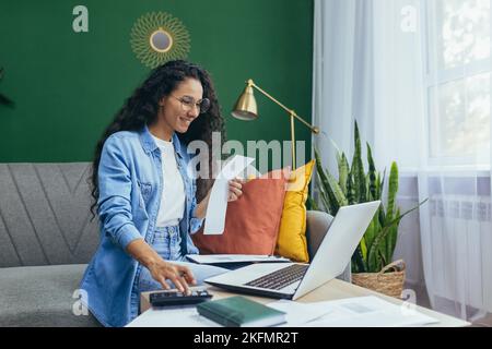 Remote work. Young beautiful Latin American businesswoman, accountant, entrepreneur works at home online. He sits on the sofa with a laptop, documents, Stock Photo